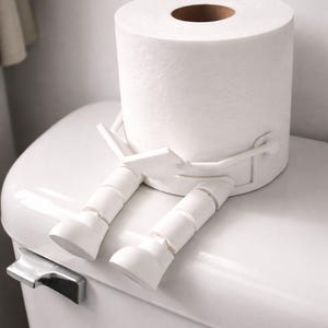 May include: A white toilet paper roll with a decorative white plastic leg attachment, sitting on a white toilet. The toilet paper roll has a brown center and is designed to look like a person sitting down.