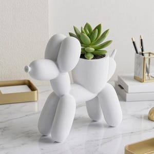 Puede incluir: Una maceta blanca con forma de perro, que se asemeja a un animal de globo, contiene una suculenta verde. La maceta está sobre una superficie de mármol, con artículos de oficina en el fondo. Un objeto decorativo único.
