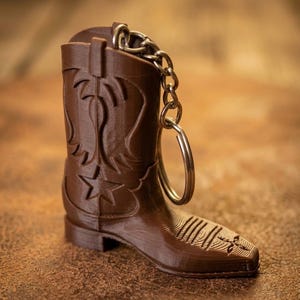 Könnte beinhalten: Ein brauner Cowboy-Stiefel-Schlüsselanhänger mit einer silbernen Kette und einem Ring. Der Stiefel hat dekorative Nähte und ein Stern-Design. Der Schlüsselanhänger besteht aus einem festen Material und ist eine Miniatur-Replik eines Cowboy-Stiefels.