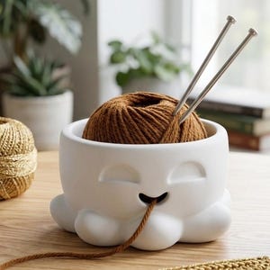 May include: A white ceramic yarn bowl shaped like an octopus with a brown yarn ball inside. Two silver knitting needles are inserted into the yarn. Other yarn balls in blue, gold, and purple are nearby, with plants in the background.