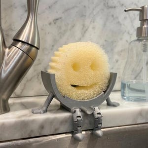 May include: A yellow sponge with a smiling face sits in a gray, 3D-printed holder with legs, designed to hang over a sink. A stainless steel faucet and soap dispenser are also visible.