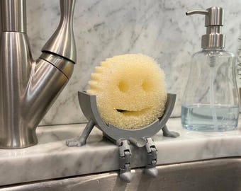 May include: A yellow sponge with a smiling face sits in a gray, 3D-printed holder with legs, designed to hang over a sink. A stainless steel faucet and soap dispenser are also visible.
