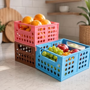 May include: A stack of three rectangular plastic storage crates with a perforated grid design. The top crate is pink and filled with oranges, the middle crate is brown, and the bottom front crate is light blue and filled with grapes and apples.