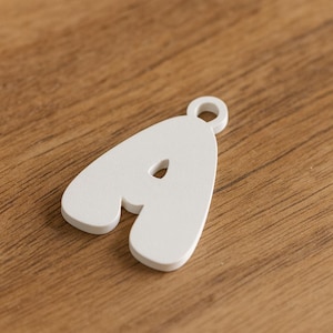 Puede incluir: Un charm de madera blanco con la letra "A", con un pequeño agujero en la parte superior para colgarlo. La letra está sobre una superficie de madera.