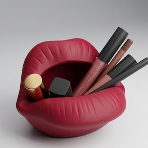 May include: A red lip-shaped container holding various makeup items. The container has a matte finish and is filled with a gold lipstick tube, a black lipstick tube, and several lip glosses and lip liners. The background is a solid, neutral color.