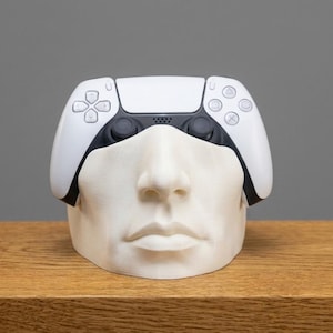 May include: A white and black video game controller resting on a white, sculpted human face stand. The controller features a directional pad, two analog sticks, and four action buttons with geometric symbols. The face sculpture is matte white.