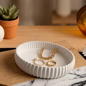 Op de afbeelding: Een witte, ovale sieradenbak met een geribbeld ontwerp, met een gouden kettingarmband en twee ringen. Een kleine potplant en decoratieve items staan op de achtergrond. De bak staat op een houten oppervlak.
