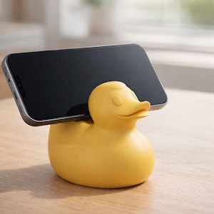 May include: A yellow duck-shaped phone stand holding a black smartphone. The stand is made of a smooth material and has a detailed duck design. The phone is positioned horizontally, resting on the duck's back.