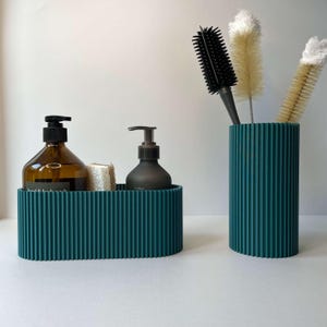 May include: Two teal ribbed containers, one oval and one cylindrical, hold bathroom essentials. The oval container holds two bottles of liquid soap and a small bar of soap. The cylindrical container holds a black hairbrush and two white bottle cleaning brushes.