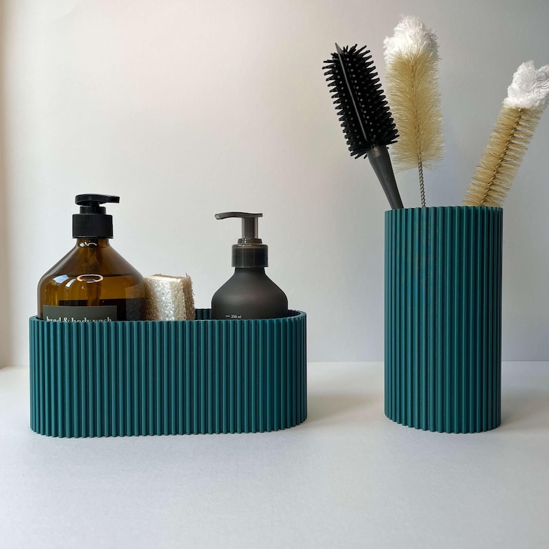 May include: Two teal ribbed containers, one oval and one cylindrical, hold bathroom essentials. The oval container holds two bottles of liquid soap and a small bar of soap. The cylindrical container holds a black hairbrush and two white bottle cleaning brushes.