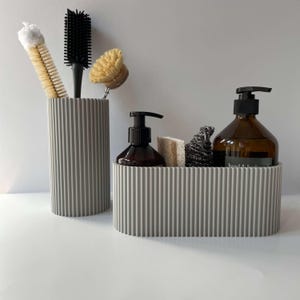 May include: Two gray ribbed containers for organizing bathroom supplies. The larger container holds a brown glass bottle with a pump dispenser, a sponge, and a black scrub brush. The smaller container holds a variety of cleaning brushes.