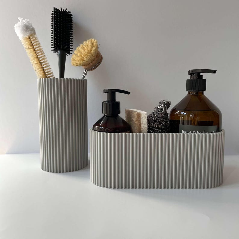 May include: Two gray ribbed containers for organizing bathroom supplies. The larger container holds a brown glass bottle with a pump dispenser, a sponge, and a black scrub brush. The smaller container holds a variety of cleaning brushes.