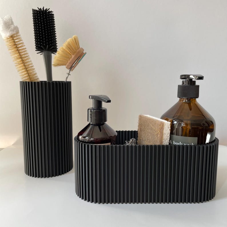 May include: Two black ribbed containers, one tall and one wide, hold various bathroom items. The tall container holds a black and white brush and a white and brown brush. The wide container holds a brown glass bottle with a pump dispenser, a brown sponge, and a clear glass bottle with a pump dispenser. The clear bottle has the text "& body wash" on the label.