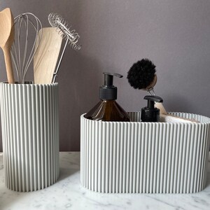 May include: Two gray ribbed containers, one tall and one wide, hold kitchen utensils and cleaning supplies. The tall container holds a wooden spoon, a whisk, and a wooden spatula. The wide container holds a bottle of liquid soap, a small bottle of liquid, and a black scrub brush.
