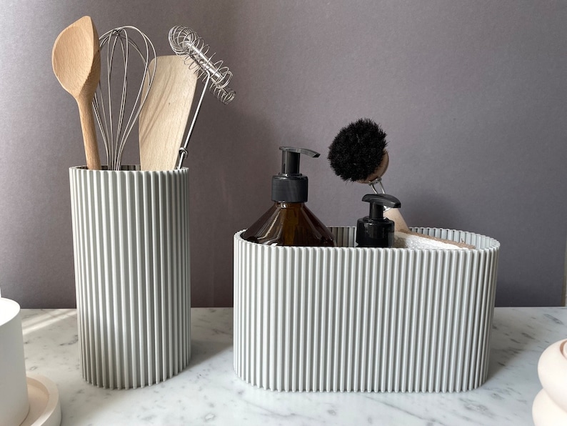 May include: Two gray ribbed containers, one tall and one wide, hold kitchen utensils and cleaning supplies. The tall container holds a wooden spoon, a whisk, and a wooden spatula. The wide container holds a bottle of liquid soap, a small bottle of liquid, and a black scrub brush.