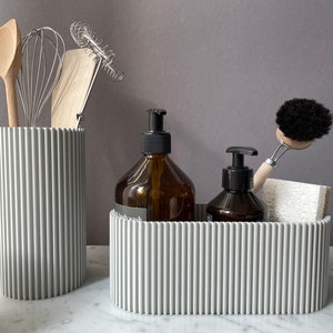 May include: Two gray ribbed containers, one tall and one wide, hold kitchen and bathroom supplies. The tall container holds wooden utensils and a whisk. The wide container holds two amber glass bottles with pumps, a black scrub brush with a wooden handle, and a white sponge.