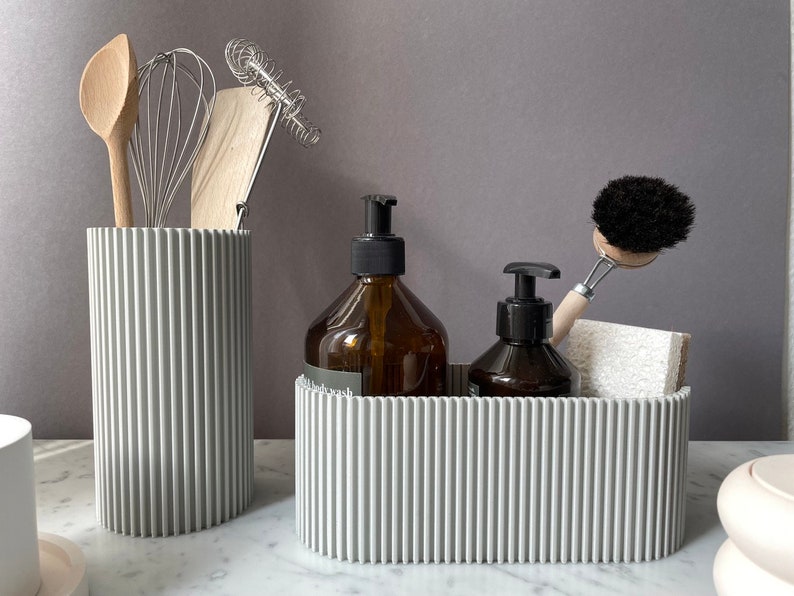 May include: Two gray ribbed containers, one tall and one wide, hold kitchen and bathroom supplies. The tall container holds wooden utensils and a whisk. The wide container holds two amber glass bottles with pumps, a black scrub brush with a wooden handle, and a white sponge.