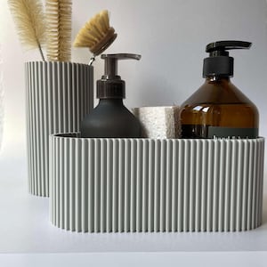 Könnte beinhalten: Ein grauer, gerippter Badezimmer-Organizer mit zwei grauen, gerippten Behältern, einer schwarzen Pumpflasche und einer braunen Glasflasche mit einem Pumpspender. Die braune Flasche trägt den Text "hand & body".