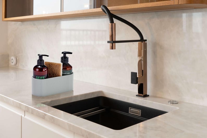 Moderner Spülbecken Organizer in Hellgrau, der mittels neustem 3D Druck in Deutschland gefertigt wird. Der Spül Organizer ist im Scandi Stil gehalten. Er ist minimalistisch designt und hat eine feine Rillenstruktur. Der Design Organizer ist befüllt mit Handseife, Spülschwamm sowie Spülmittel. Der stylische Küchen Organizer steht in einer modernen aufgeräumten Küche auf einer Marmorarbeitsplatte neben einem schwarzen Spülbecken mit Messing Wasserhahn. Utensilienhalter für Ordnung in der Küche.