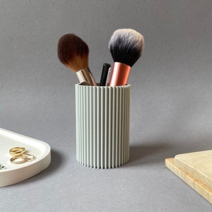 May include: A light gray, ribbed cylindrical container holding makeup brushes. The brushes have brown and black bristles with silver and copper-colored handles. A white tray with gold rings is in the background.