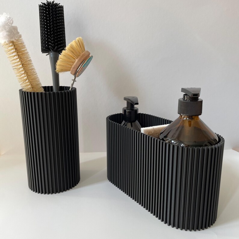 May include: Two black ribbed containers for bathroom storage. One container holds a bottle of soap and a bar of soap. The other container holds a variety of cleaning brushes.