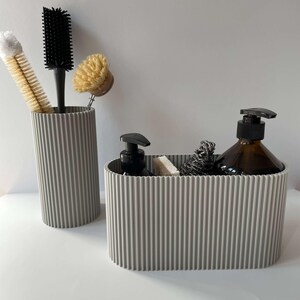 May include: Two gray ribbed silicone containers for organizing bathroom supplies. One container is tall and cylindrical, holding a black brush and a yellow brush. The other container is oval and holds a brown soap dispenser, a white sponge, and a gray scrub brush.