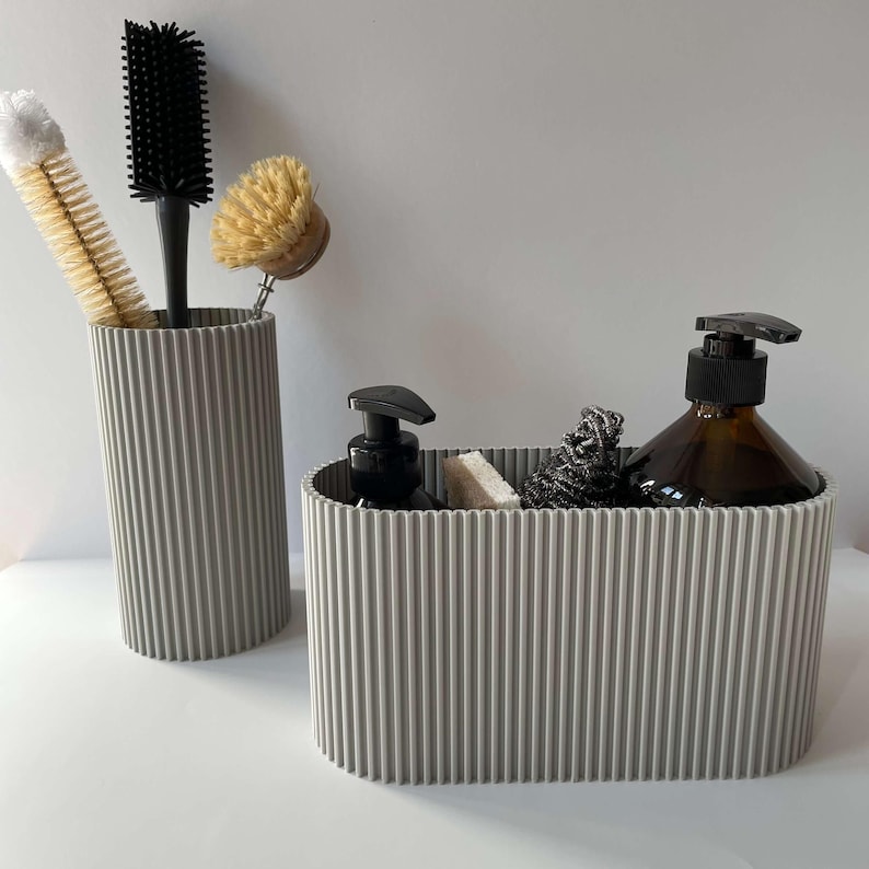 May include: Two gray ribbed silicone containers for organizing bathroom supplies. One container is tall and cylindrical, holding a black brush and a yellow brush. The other container is oval and holds a brown soap dispenser, a white sponge, and a gray scrub brush.