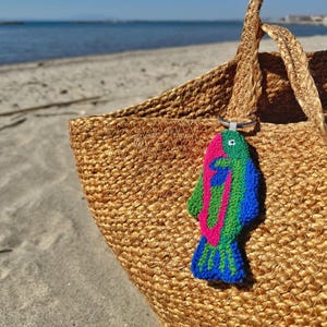 May include: A woven straw tote bag with a colorful fish-shaped keychain. The keychain features a vibrant design with pink, green, and blue yarn. The bag is on a sandy beach with the ocean in the background.