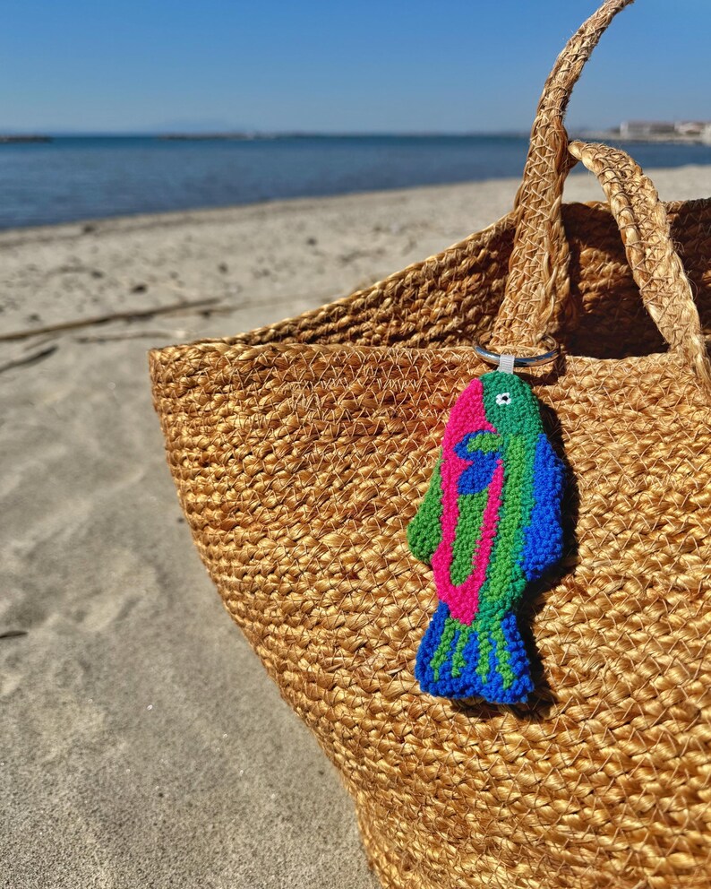 May include: A woven straw tote bag with a colorful fish-shaped keychain. The keychain features a vibrant design with pink, green, and blue yarn. The bag is on a sandy beach with the ocean in the background.