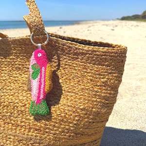 May include: A woven straw tote bag with a colorful fish-shaped keychain. The keychain features pink, green, and yellow details. The bag is on a sandy beach with the ocean in the background.