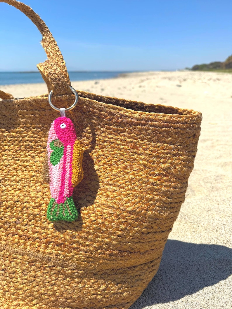 May include: A woven straw tote bag with a colorful fish-shaped keychain. The keychain features pink, green, and yellow details. The bag is on a sandy beach with the ocean in the background.