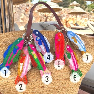 May include: A straw tote bag with a brown leather handle, adorned with seven colorful, fish-shaped keychains. The keychains are intricately designed with various colors, including pink, green, blue, and orange, and are attached to the bag with metal rings.