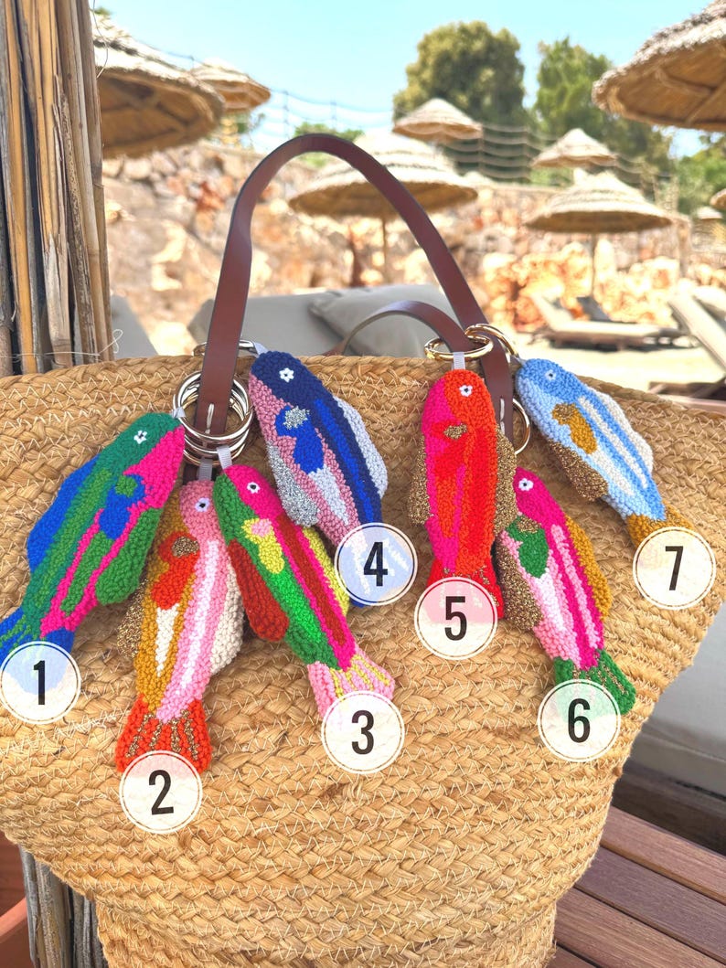 May include: A straw tote bag with a brown leather handle, adorned with seven colorful, fish-shaped keychains. The keychains are intricately designed with various colors, including pink, green, blue, and orange, and are attached to the bag with metal rings.