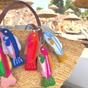 May include: A collection of colorful, handmade fish keychains. Each keychain features a unique design with vibrant hues of pink, blue, red, and yellow. The keychains are attached to a brown leather bag handle, resting on a woven straw bag.