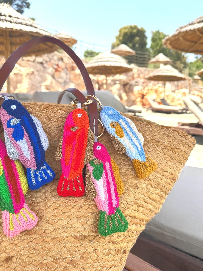 May include: A collection of colorful, handmade fish keychains. Each keychain features a unique design with vibrant hues of pink, blue, red, and yellow. The keychains are attached to a brown leather bag handle, resting on a woven straw bag.