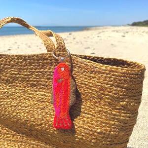 May include: A woven straw tote bag with a colorful fish-shaped keychain. The keychain is red, orange, and pink. The bag is sitting on a sandy beach with the ocean in the background.