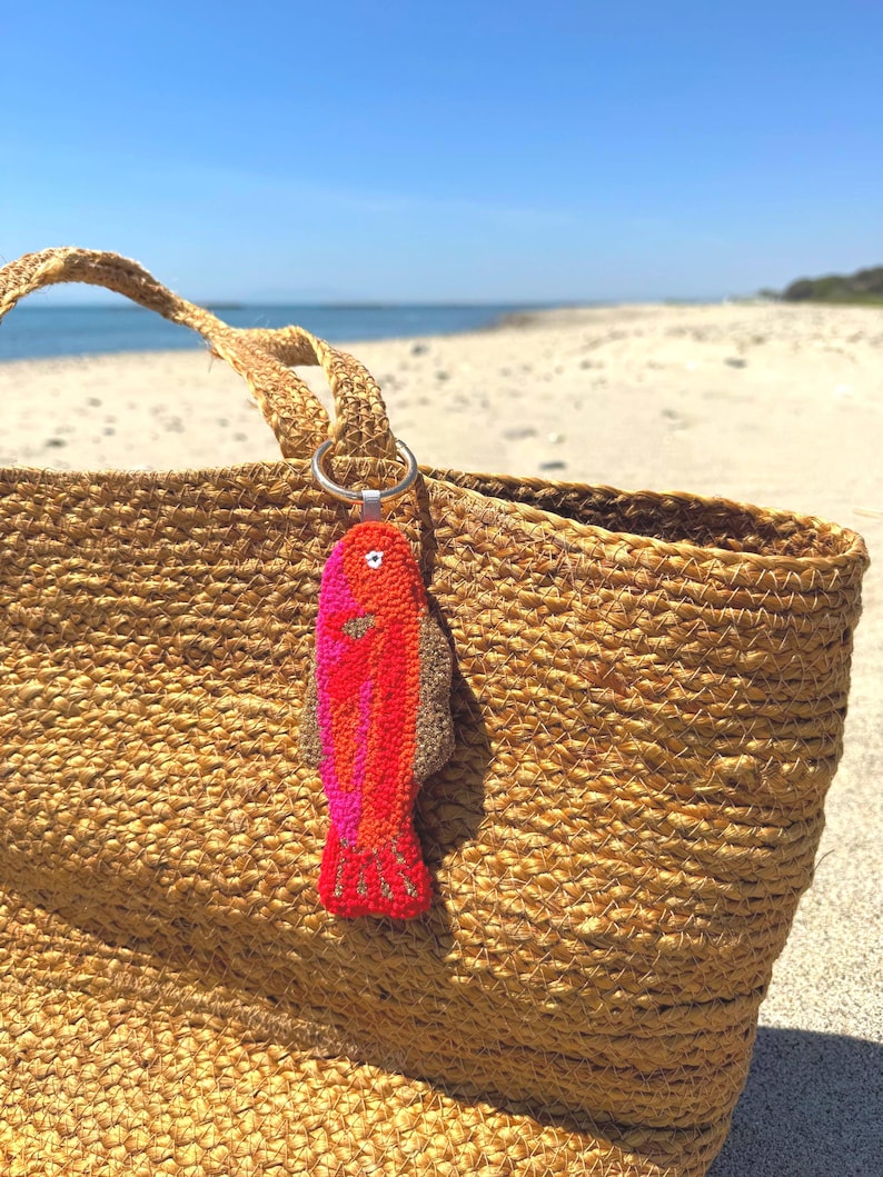 May include: A woven straw tote bag with a colorful fish-shaped keychain. The keychain is red, orange, and pink. The bag is sitting on a sandy beach with the ocean in the background.