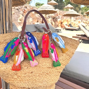 May include: A woven straw tote bag with a brown leather handle, decorated with vibrant, embroidered fish keyrings. The fish are in shades of pink, blue, green, and orange, attached to silver keyrings. The bag is on a wooden surface.