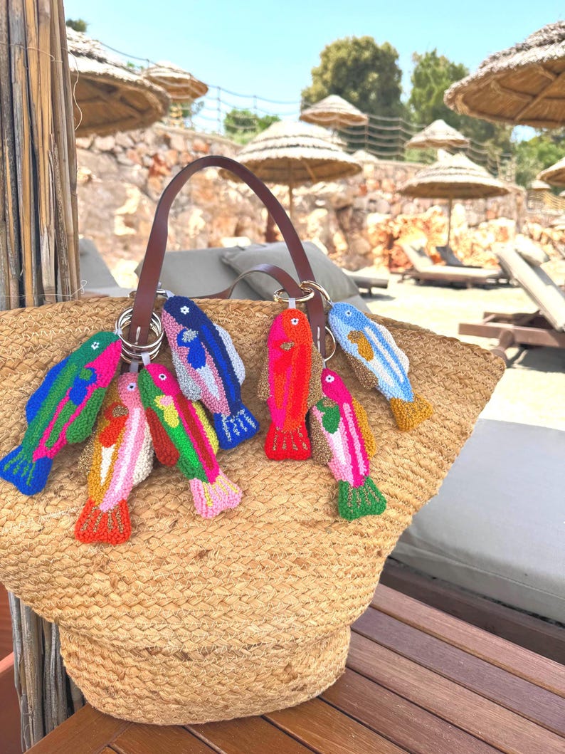 May include: A woven straw tote bag with a brown leather handle, adorned with colorful, embroidered fish keychains. The fish feature various colors like pink, blue, green, and orange, and are attached to silver keyrings.