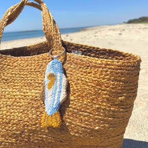 May include: A woven straw tote bag with a blue, white, and yellow fish-shaped keychain attached to the handle. The bag is tan and sits on a sandy beach with the ocean in the background.