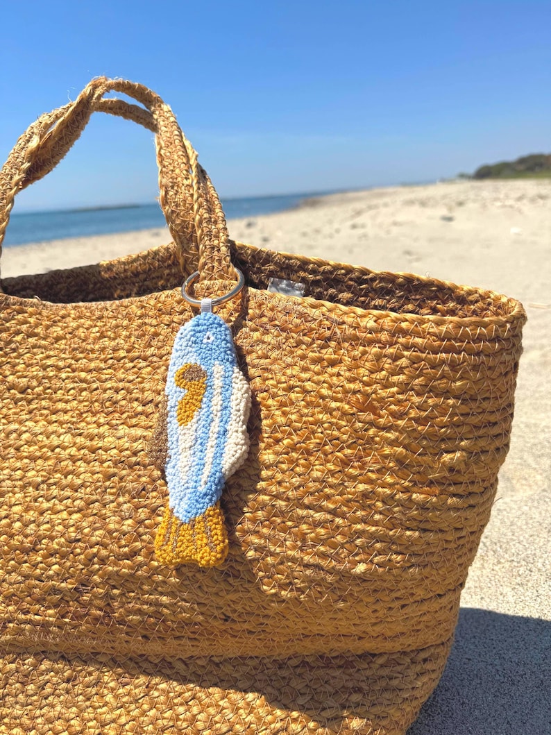 May include: A woven straw tote bag with a blue, white, and yellow fish-shaped keychain attached to the handle. The bag is tan and sits on a sandy beach with the ocean in the background.
