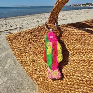 May include: A woven jute tote bag with a colorful fish-shaped keychain. The keychain features pink, green, and yellow details. The bag is on a sandy beach with the ocean in the background.