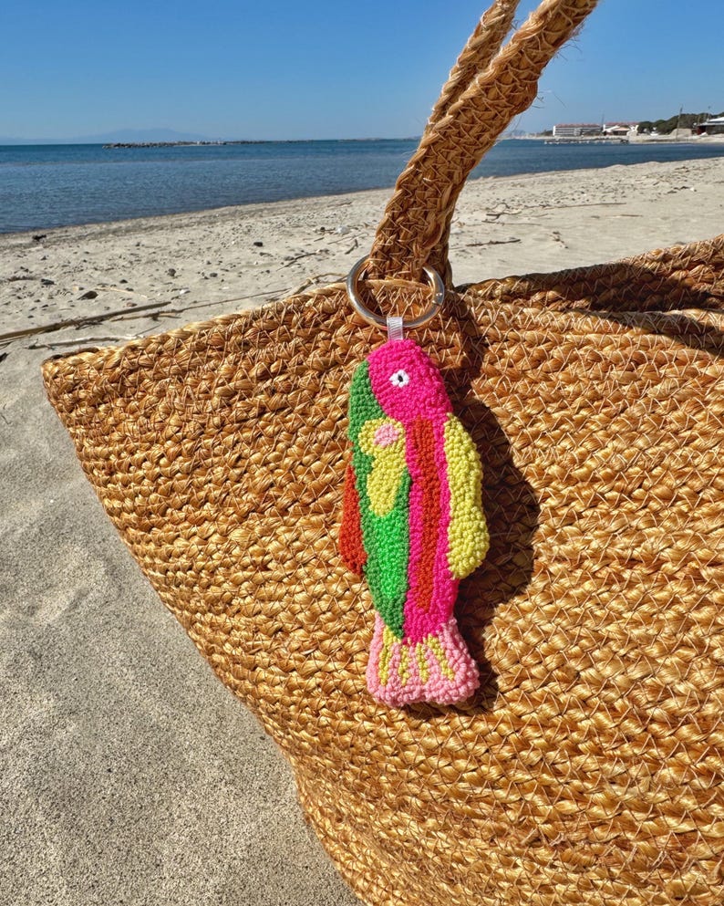 May include: A woven jute tote bag with a colorful fish-shaped keychain. The keychain features pink, green, and yellow details. The bag is on a sandy beach with the ocean in the background.