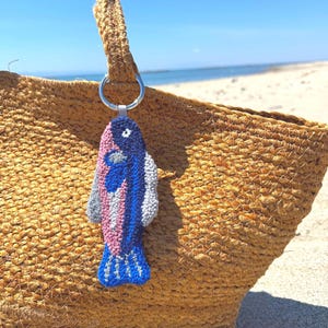May include: A colorful fish-shaped keychain with blue, pink, and gray details hangs from a straw tote bag. The keychain has a silver ring and is set against a beach backdrop with a blue sky and ocean.