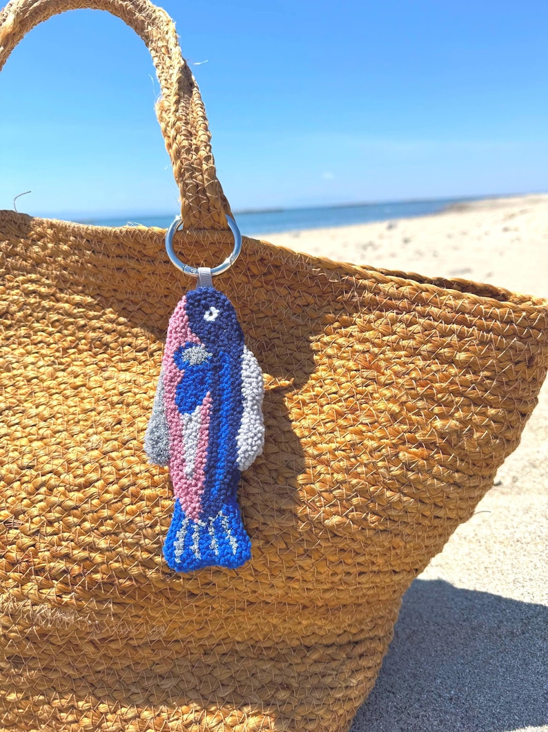 May include: A colorful fish-shaped keychain with blue, pink, and gray details hangs from a straw tote bag. The keychain has a silver ring and is set against a beach backdrop with a blue sky and ocean.
