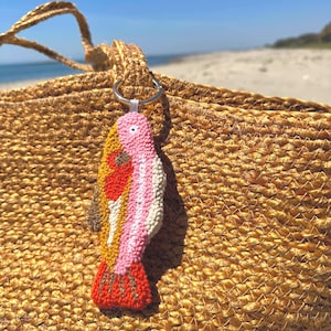 May include: A colorful, handmade fish-shaped keychain with a silver ring. The fish features a textured design with pink, white, yellow, orange, and brown details. The keychain is attached to a woven straw bag, with a beach and blue sky in the background.