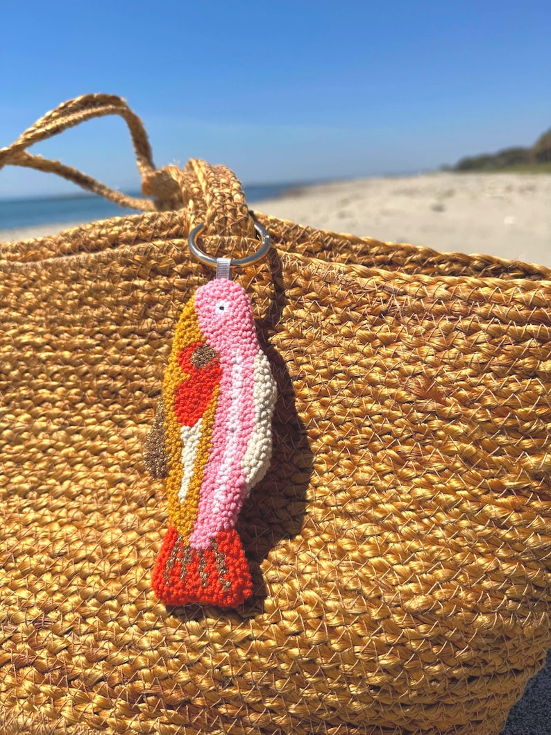 May include: A colorful, handmade fish-shaped keychain with a silver ring. The fish features a textured design with pink, white, yellow, orange, and brown details. The keychain is attached to a woven straw bag, with a beach and blue sky in the background.