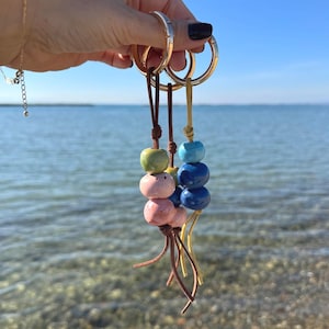 May include: Keyrings with ceramic beads in green, pink, and blue. Each keyring has a leather cord and a gold-coloured metal ring. The background shows a clear blue sky and sea. The beads are in various sizes.