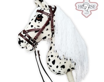Puede incluir: Una cabeza de caballo de peluche blanca y negra con una brida y riendas de cuero marrón. El caballo tiene una crin y una cola blancas. El caballo está montado en un palo de madera.