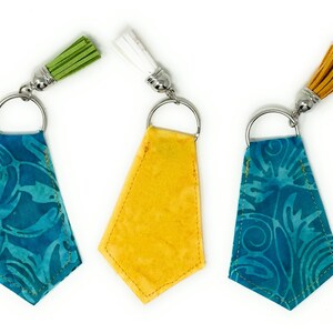 Op de afbeelding: Drie stoffen sleutelhangers met verschillende stoffen en kwastjes. De sleutelhangers zijn turquoise met een wit patroon, geel en turquoise met een blauw patroon.